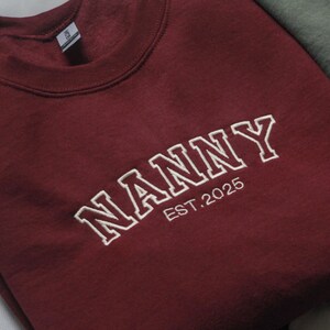 K&ouml;nnte beinhalten: Ein weinrotes Sweatshirt mit dem wei&szlig;en gestickten Wort "NANNY" und der Jahreszahl "EST.2025". Das Sweatshirt mit Rundhalsausschnitt ist gefaltet und wirkt weich. Das Kleidungsst&uuml;ck ist wahrscheinlich aus einem bequemen Material.