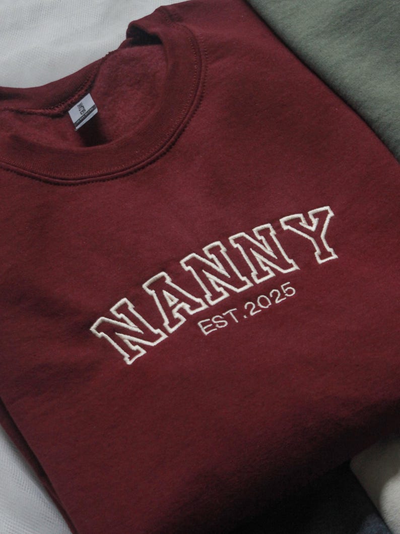 K&ouml;nnte beinhalten: Ein weinrotes Sweatshirt mit dem wei&szlig;en gestickten Wort "NANNY" und der Jahreszahl "EST.2025". Das Sweatshirt mit Rundhalsausschnitt ist gefaltet und wirkt weich. Das Kleidungsst&uuml;ck ist wahrscheinlich aus einem bequemen Material.