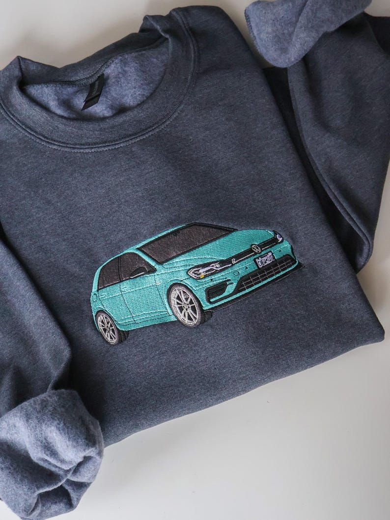 May include: A dark grey sweatshirt featuring an embroidered teal car. The car has silver wheels and a black roof. The sweatshirt has a crew neck and long sleeves. The car's license plate is visible.
