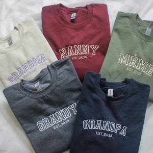 K&ouml;nnte beinhalten: F&uuml;nf Sweatshirts in Beige, Burgunderrot, Olivgr&uuml;n, Anthrazitgrau und Marineblau. Jedes Sweatshirt hat einen gestickten wei&szlig;en Namen: "Grandma", "Nanny", "Meme", "Grandy" und "Grandpa", darunter "EST.2025". Verschiedene Farben und Stile.