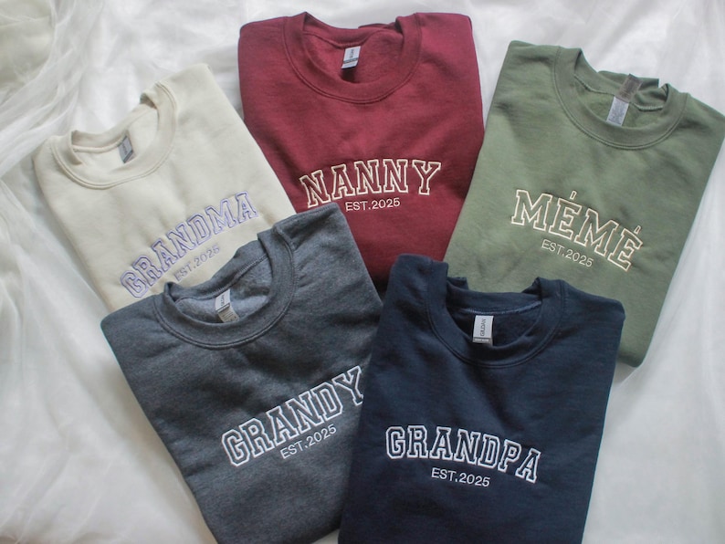 K&ouml;nnte beinhalten: F&uuml;nf Sweatshirts in Beige, Burgunderrot, Olivgr&uuml;n, Anthrazitgrau und Marineblau. Jedes Sweatshirt hat einen gestickten wei&szlig;en Namen: "Grandma", "Nanny", "Meme", "Grandy" und "Grandpa", darunter "EST.2025". Verschiedene Farben und Stile.