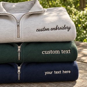 Peut inclure: Une pile de trois sweat-shirts à fermeture éclair quart de longueur en gris clair, vert foncé et bleu marine. Le sweat gris porte le texte brodé noir custom embroidery, le vert custom text et le bleu marine your text here.