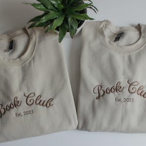 May include: Two beige crewneck sweatshirts embroidered with "Book Club Est. 2021" in a brown script font. A small green plant sits between the sweatshirts. The sweatshirts are folded neatly on a white surface.