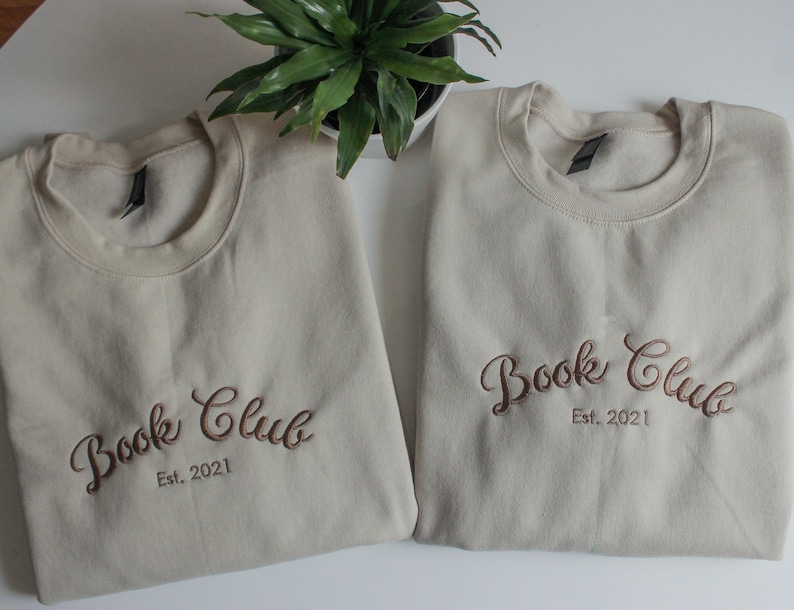 May include: Two beige crewneck sweatshirts embroidered with "Book Club Est. 2021" in a brown script font. A small green plant sits between the sweatshirts. The sweatshirts are folded neatly on a white surface.