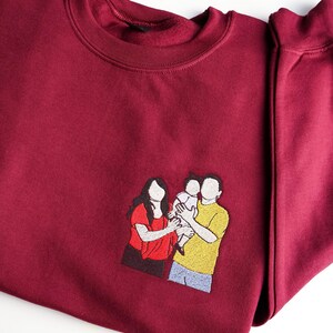 Puede incluir: Una sudadera burdeos con un retrato familiar bordado. El retrato representa a una madre, un padre y un ni&ntilde;o en un estilo minimalista. La madre lleva una camisa roja, el padre una camisa amarilla y el ni&ntilde;o va de blanco.