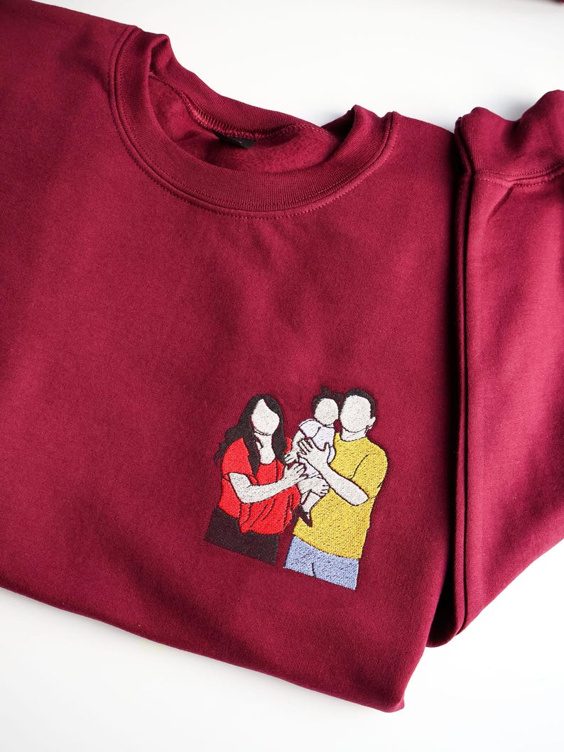 Puede incluir: Una sudadera burdeos con un retrato familiar bordado. El retrato representa a una madre, un padre y un ni&ntilde;o en un estilo minimalista. La madre lleva una camisa roja, el padre una camisa amarilla y el ni&ntilde;o va de blanco.