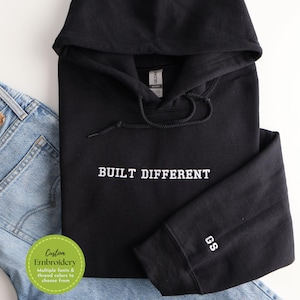 May include: Black hoodie with the text "BUILT DIFFERENT" in white. The hoodie has a drawstring and a hood. The initials "GS" are embroidered on the sleeve. Light blue denim jeans are visible in the background. A green circle with text is in the lower left corner.