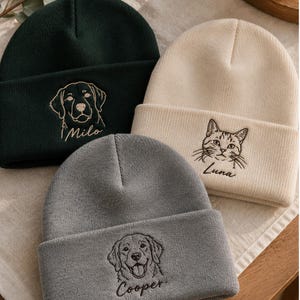 May include: Three knit beanies in dark green, cream, and grey. Each beanie features a detailed embroidered animal portrait and a name: a dog labelled Milo, a cat labelled Luna, and a dog labelled Cooper. The hats have a folded cuff design.