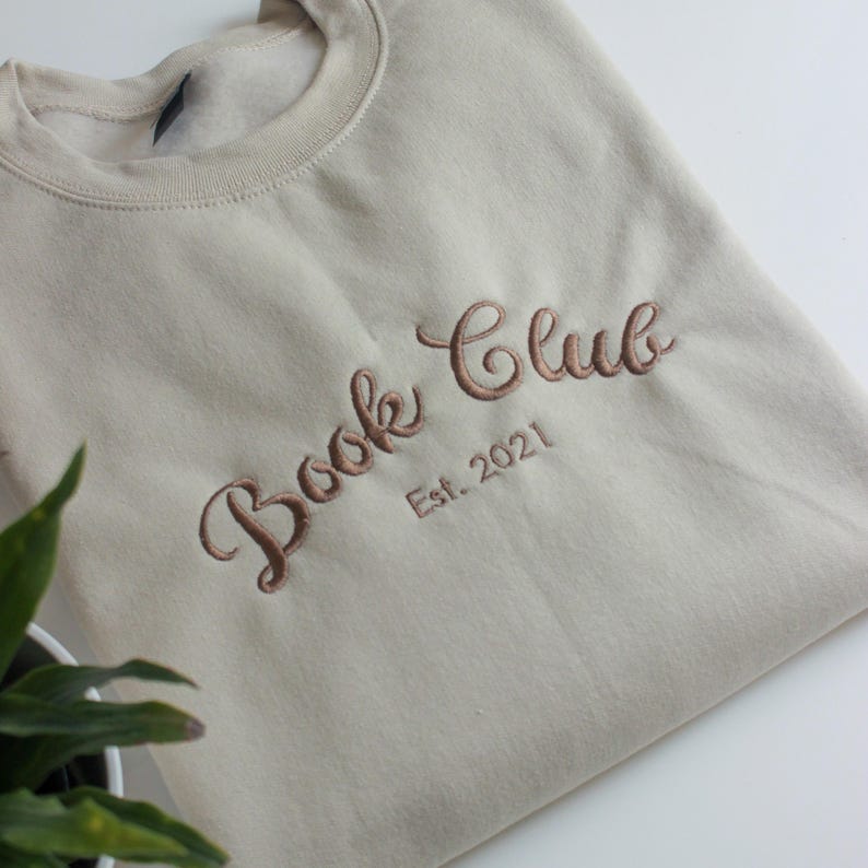 May include: A beige sweatshirt with the embroidered text "Book Club Est. 2021" in a brown script font. The sweatshirt is made of a soft material and has a crew neck.