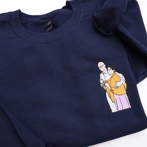 Puede incluir: Sudadera azul marino con un dise&ntilde;o bordado de dos personas abraz&aacute;ndose. Las figuras est&aacute;n representadas en un estilo minimalista, con una persona con gafas y camisa blanca, y la otra con un chal amarillo y falda rosa.