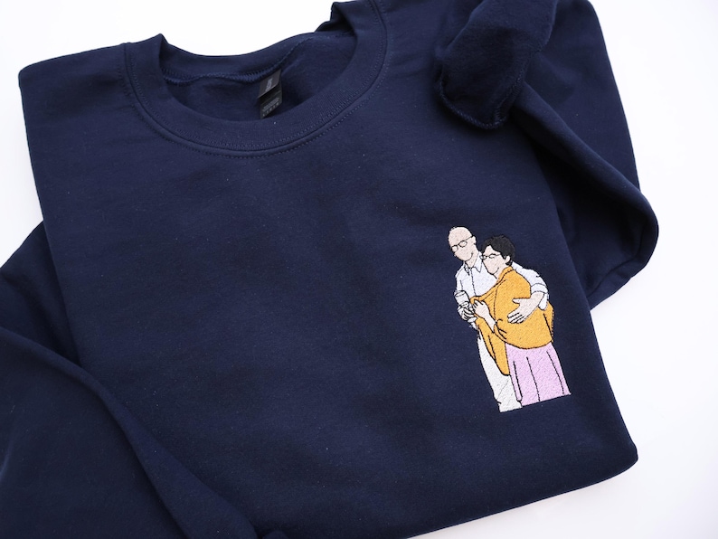 Puede incluir: Sudadera azul marino con un dise&ntilde;o bordado de dos personas abraz&aacute;ndose. Las figuras est&aacute;n representadas en un estilo minimalista, con una persona con gafas y camisa blanca, y la otra con un chal amarillo y falda rosa.