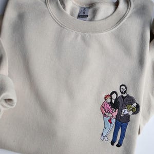 Puede incluir: Sudadera beige con un retrato familiar bordado. El retrato representa a una pareja con dos hijos, uno con una chaqueta rosa y el otro en brazos del padre. La sudadera tiene cuello redondo y mangas largas.