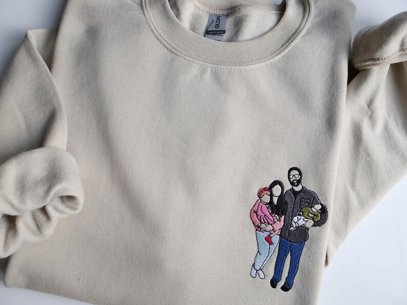 Puede incluir: Sudadera beige con un retrato familiar bordado. El retrato representa a una pareja con dos hijos, uno con una chaqueta rosa y el otro en brazos del padre. La sudadera tiene cuello redondo y mangas largas.