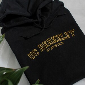 K&ouml;nnte beinhalten: Schwarzer Kapuzenpullover mit goldfarben gesticktem Schriftzug "UC BERKELEY STATISTICS". Der Pullover hat eine Kapuze mit Kordelzug und ist gefaltet. Der Text ist in einem klassischen College-Stil gehalten.