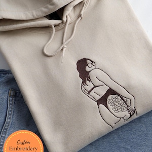 May include: A beige hooded sweatshirt with a drawstring. It has an embroidered design of a person in a swimsuit. The text "Custom Embroidery Design shared for approval" is in a circle on the bottom left.