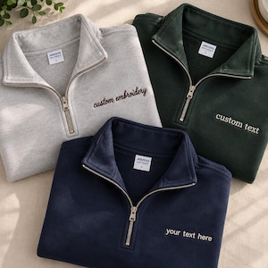 Könnte beinhalten: Drei Sweatshirts mit kurzem Reißverschluss in Hellgrau, Dunkelgrün und Marineblau. Jedes verfügt über einen Metallreißverschluss, ein Jerzees-Markenetikett und gestickten Text auf der Brust, der jeweils individuell angepasst ist.