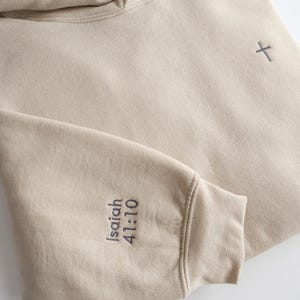 May include: A beige hoodie with a drawstring hood and a small cross embroidered on the chest. The left sleeve cuff features the embroidered text "Isaiah 41:10" in a dark gray color. The hoodie is made of a soft, comfortable material.