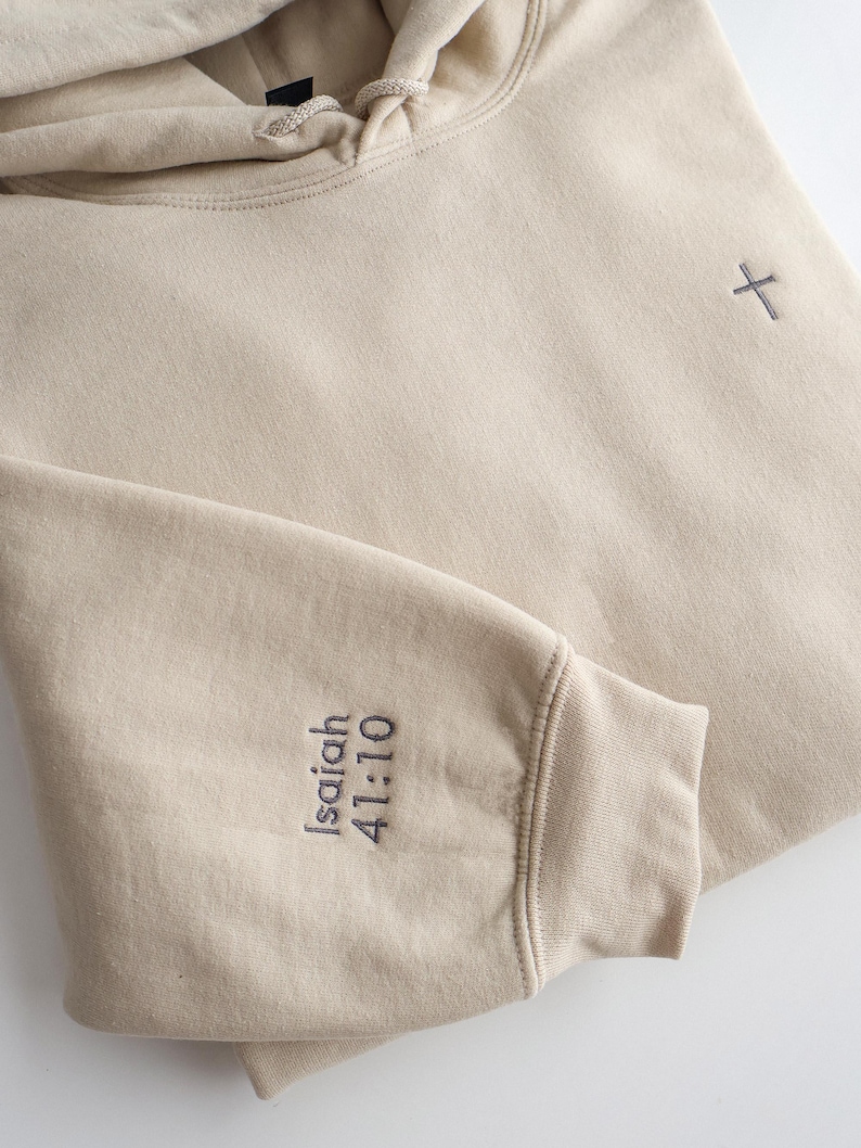 May include: A beige hoodie with a drawstring hood and a small cross embroidered on the chest. The left sleeve cuff features the embroidered text "Isaiah 41:10" in a dark gray color. The hoodie is made of a soft, comfortable material.