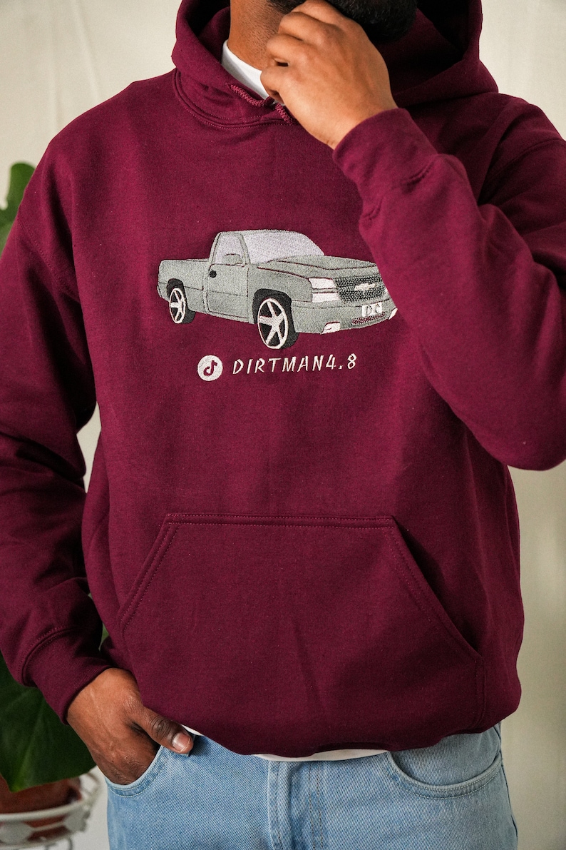 May include: Burgundy hoodie featuring a grey truck graphic. The truck design is embroidered on the front, with the text "Dirtman4.8" and a TikTok logo. The hoodie has a front pocket and a hood.