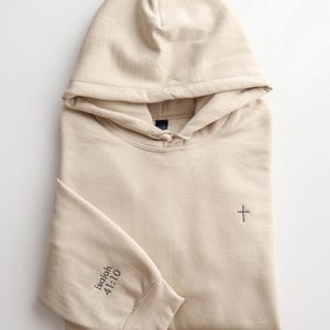 May include: A beige hoodie with a hood and long sleeves. A small gray cross is embroidered on the chest. The left sleeve cuff has the text "Isaiah 41:10" printed in gray. The hoodie is folded neatly on a white surface.