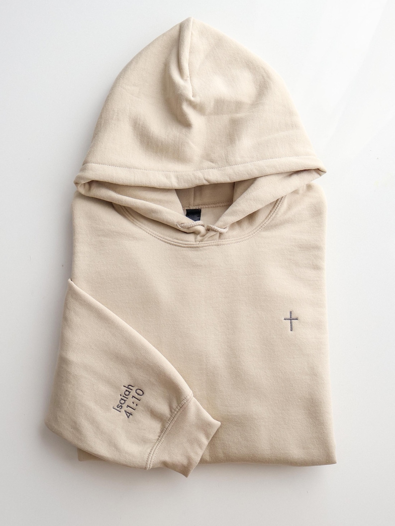 May include: A beige hoodie with a hood and long sleeves. A small gray cross is embroidered on the chest. The left sleeve cuff has the text "Isaiah 41:10" printed in gray. The hoodie is folded neatly on a white surface.