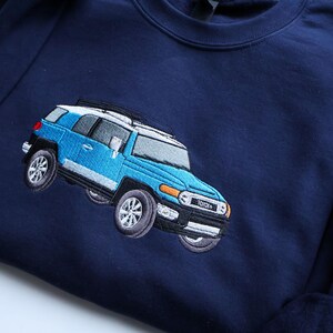 May include: Navy blue sweatshirt with an embroidered light blue Toyota FJ Cruiser. The vehicle has a white roof, silver accents, and detailed wheels. The sweatshirt is made of a soft material.