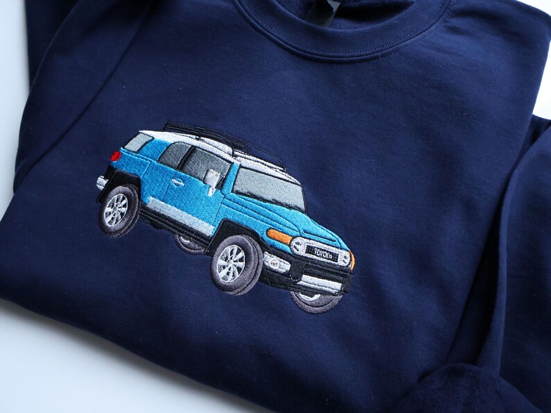 May include: Navy blue sweatshirt with an embroidered light blue Toyota FJ Cruiser. The vehicle has a white roof, silver accents, and detailed wheels. The sweatshirt is made of a soft material.