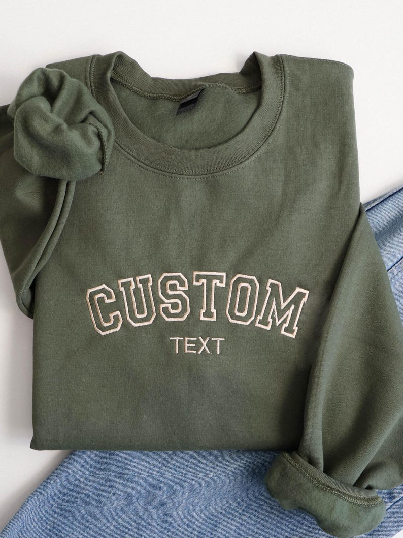 K&ouml;nnte beinhalten: Ein olivgr&uuml;nes Sweatshirt mit dem Wort "CUSTOM" in cremefarbener Schrift im College-Stil gestickt. Unter dem Wort "CUSTOM" steht das Wort "TEXT". Das Sweatshirt ist gefaltet, mit einem Scrunchie und Jeans sichtbar.
