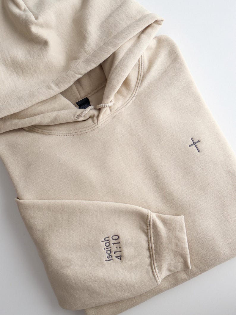 May include: A beige hoodie with a drawstring hood and a small embroidered cross on the chest. The left sleeve features the embroidered text "Isaiah 41:10". The hoodie is made of a soft material.