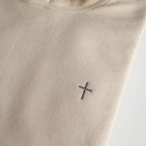 May include: A beige hoodie with a drawstring hood and a small, dark gray cross embroidered on the chest. The hoodie is made of a soft, textured fabric.