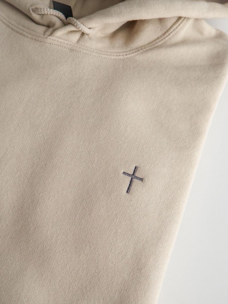 May include: A beige hoodie with a drawstring hood and a small, dark gray cross embroidered on the chest. The hoodie is made of a soft, textured fabric.