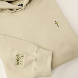 May include: A beige hoodie with a drawstring hood and a small green cross embroidered on the chest. The left sleeve cuff features the embroidered text "Isaiah 41:10". The hoodie is folded, showing the sleeve and part of the hood.