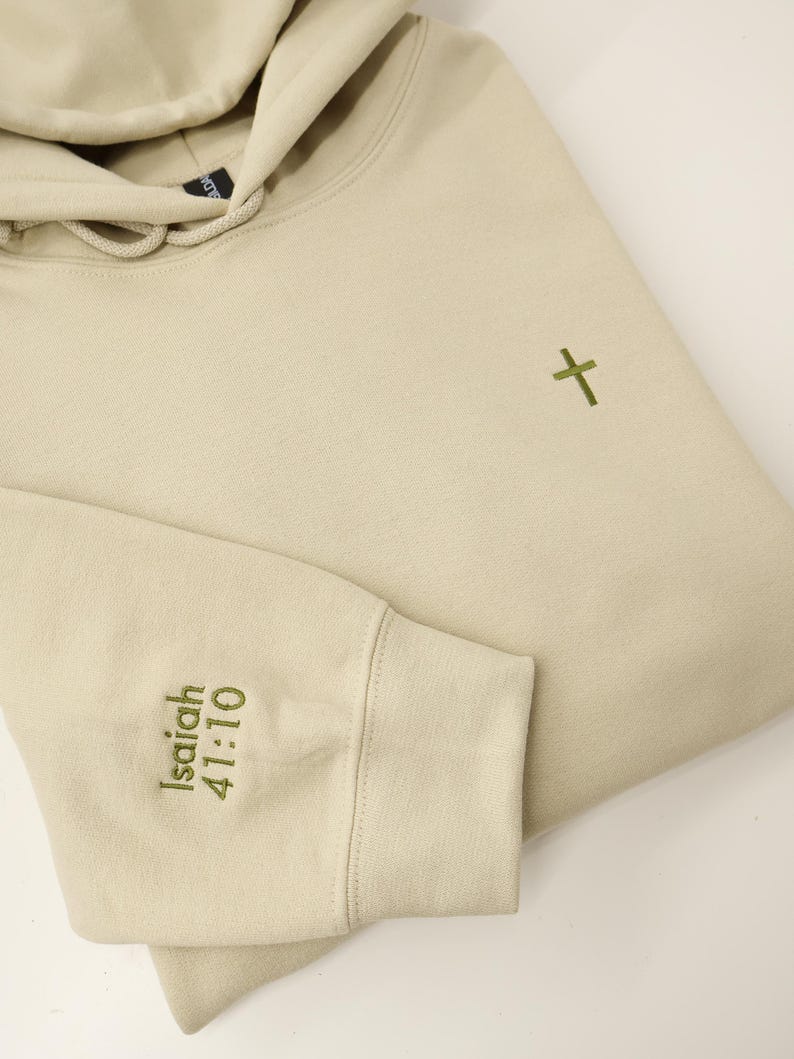 May include: A beige hoodie with a drawstring hood and a small green cross embroidered on the chest. The left sleeve cuff features the embroidered text "Isaiah 41:10". The hoodie is folded, showing the sleeve and part of the hood.