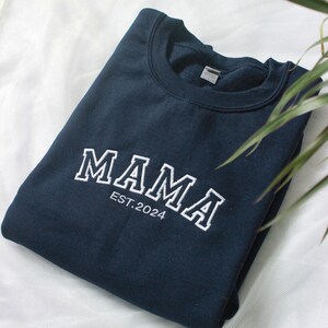 K&ouml;nnte beinhalten: Ein marineblaues Sweatshirt mit dem wei&szlig;en, gestickten Wort "MAMA" in Bogenform auf der Brust, darunter "EST.2024". Das Sweatshirt ist ordentlich gefaltet auf einer wei&szlig;en Oberfl&auml;che.