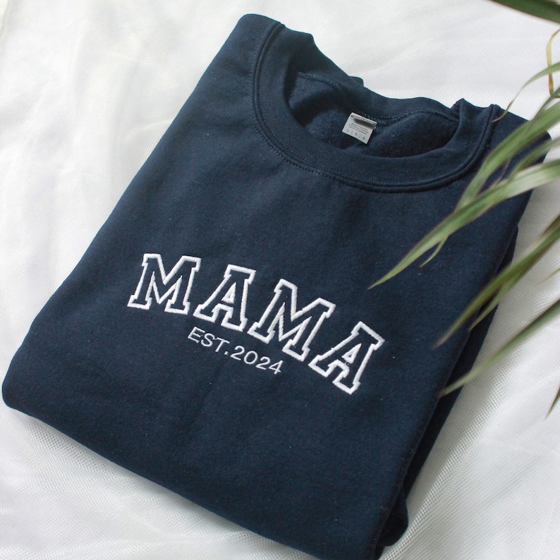 K&ouml;nnte beinhalten: Ein marineblaues Sweatshirt mit dem wei&szlig;en, gestickten Wort "MAMA" in Bogenform auf der Brust, darunter "EST.2024". Das Sweatshirt ist ordentlich gefaltet auf einer wei&szlig;en Oberfl&auml;che.