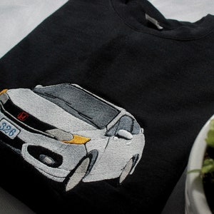 May include: Black sweatshirt with an embroidered white car design. The car has grey and yellow accents, a red emblem, and a license plate with the number "326". The sweatshirt is folded on a white surface.