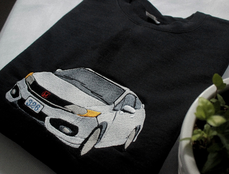 May include: Black sweatshirt with an embroidered white car design. The car has grey and yellow accents, a red emblem, and a license plate with the number "326". The sweatshirt is folded on a white surface.