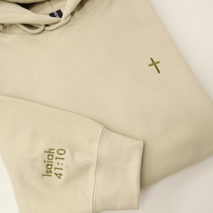Peut inclure: Un sweat à capuche beige avec un cordon de serrage et une petite croix verte brodée sur la poitrine. Le poignet de la manche gauche porte la broderie "Isaïe 41:10". Le sweat à capuche est plié, montrant la manche et une partie de la capuche.