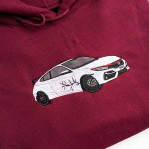 May include: Burgundy-coloured hoodie with an embroidered white car design. The car has black wheels and red detailing. The hoodie features a drawstring hood and is made of a soft material.