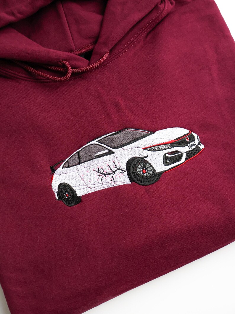 May include: Burgundy-coloured hoodie with an embroidered white car design. The car has black wheels and red detailing. The hoodie features a drawstring hood and is made of a soft material.