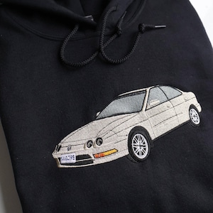 May include: Black hooded sweatshirt featuring an embroidered white car. The car has silver rims and a license plate reading "J4039E". The hood has black drawstrings.