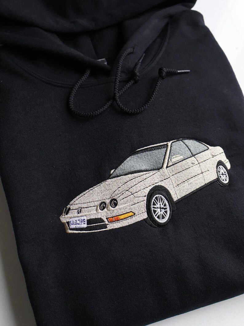May include: Black hooded sweatshirt featuring an embroidered white car. The car has silver rims and a license plate reading "J4039E". The hood has black drawstrings.