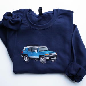 May include: A navy blue sweatshirt featuring an embroidered light blue Toyota FJ Cruiser. The vehicle has silver detailing and black tyres. The sweatshirt is folded, with the sleeves and collar visible.