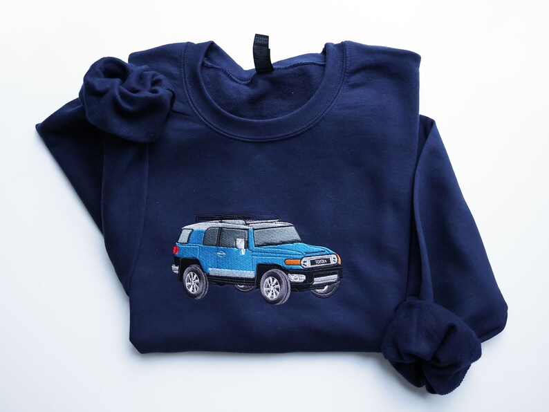 May include: A navy blue sweatshirt featuring an embroidered light blue Toyota FJ Cruiser. The vehicle has silver detailing and black tyres. The sweatshirt is folded, with the sleeves and collar visible.