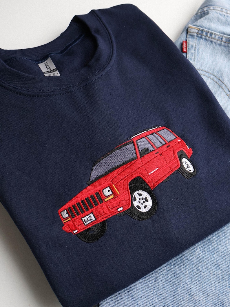 May include: A navy blue sweatshirt with a red embroidered SUV design. The SUV has black tyres and a license plate reading "LIZ". The sweatshirt is shown with blue denim jeans.