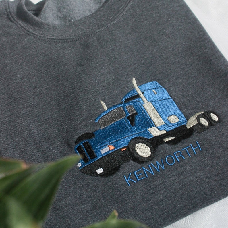 May include: Dark grey sweatshirt with a blue embroidered Kenworth lorry design. The lorry features blue, black, and white details. The word "KENWORTH" is embroidered in blue beneath the lorry.