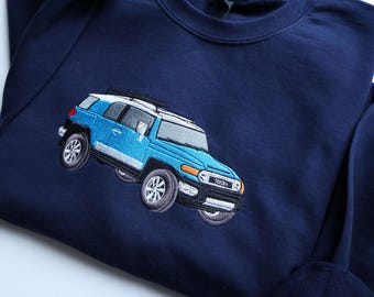 May include: Navy blue sweatshirt with an embroidered light blue Toyota FJ Cruiser. The vehicle has a white roof, silver accents, and detailed wheels. The sweatshirt is made of a soft material.