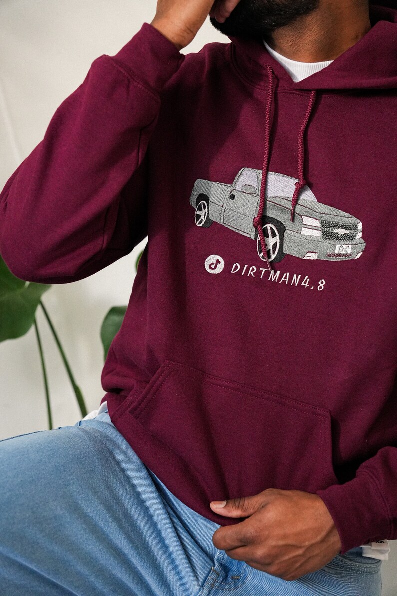 May include: Burgundy hoodie featuring a grey truck graphic and the text "DIRTMAN4.8". The hoodie has a front pocket and a drawstring hood. The person in the image is wearing blue jeans.