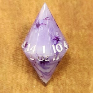 May include: A large, clear, purple, and white D20 dice with a floral design. The dice has white numbers and is made of resin. The dice is a polyhedron with 20 sides, used for tabletop games.