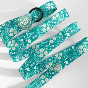 May include: A teal dog leash and collar set with a floral pattern of white, yellow, and purple flowers. The leash has a black metal clasp and a black metal ring. The collar has a round button with the text "WYLD CUB".